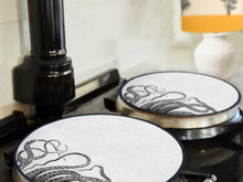 Load image into Gallery viewer, black kraken squid octocpus tenticles print on a beige linen circular aga cover with black hemming. Pictured on metal aga lid. Mustard and Gray