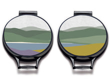 Load image into Gallery viewer, Simple landscape illustration of hills, lake and foreground of purple heather and yellow gorse printed onto neutral linen circular hob covers and hemmed in black. Pictured as aga chef&#39;s pads on aga lids with an isolated background. Aga pads from Mustard and Gray 
